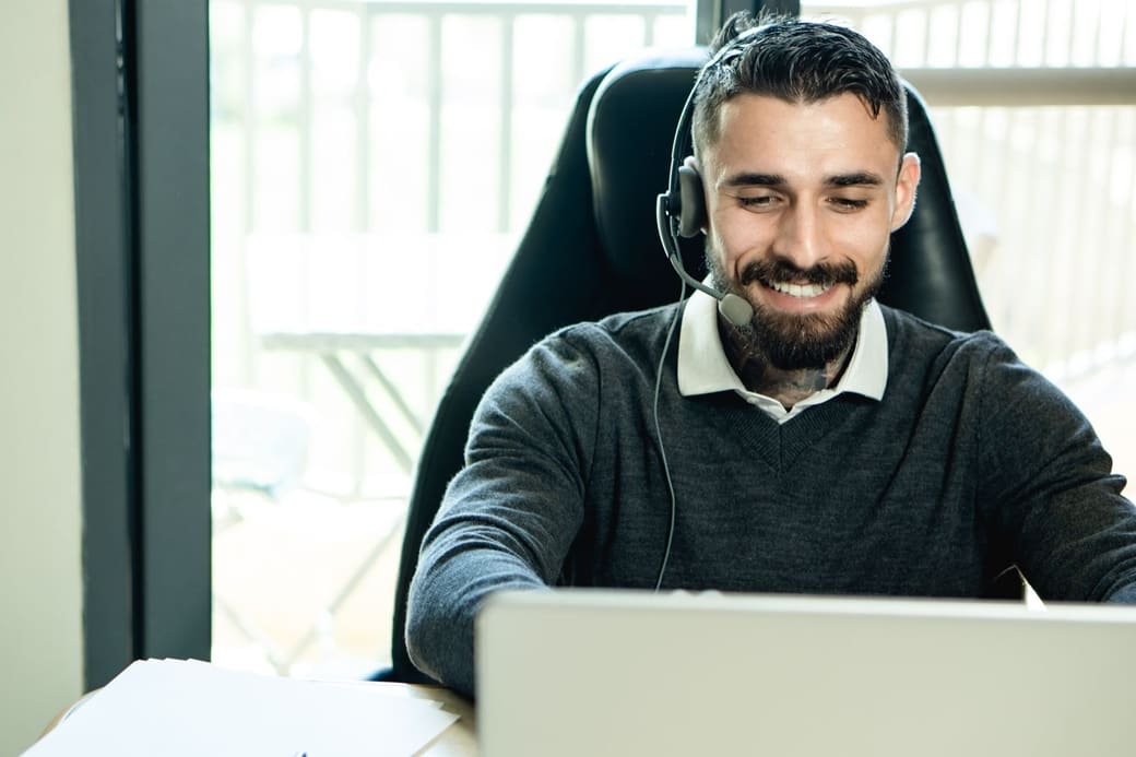 A man in a headset working on a laptop at an office desk, mid-call