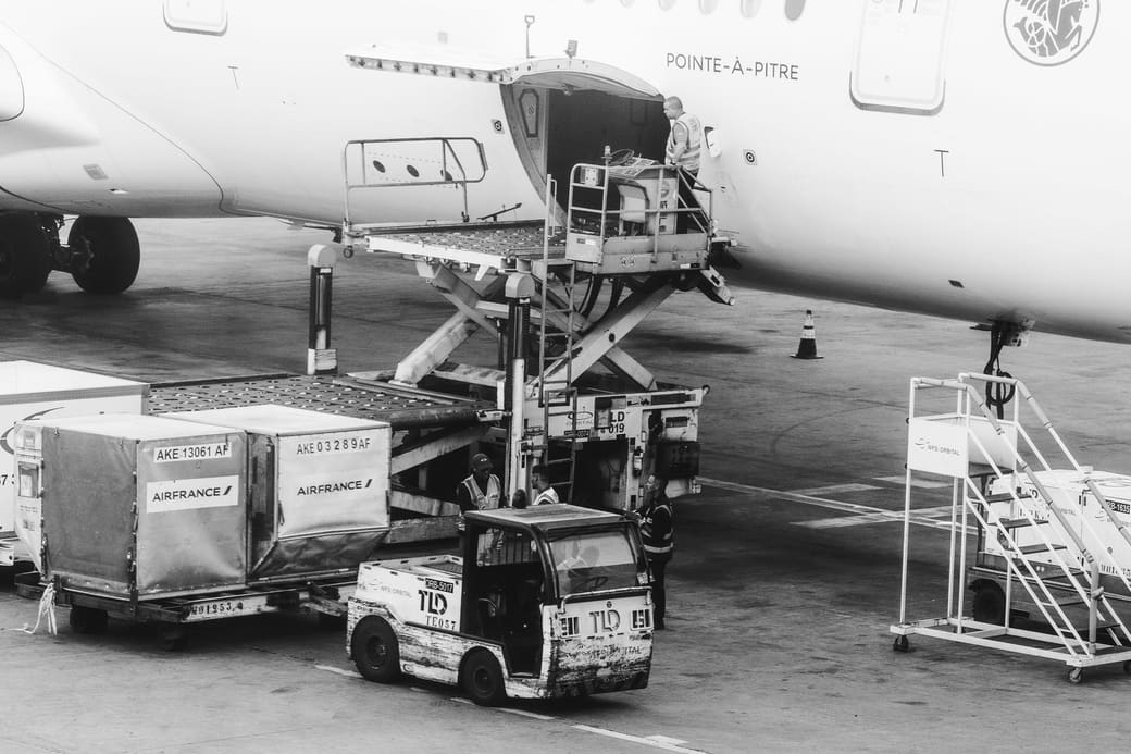 Ground crew loading air cargo containers into the hold of a passenger jet from a scissor-lift loader, in black and white