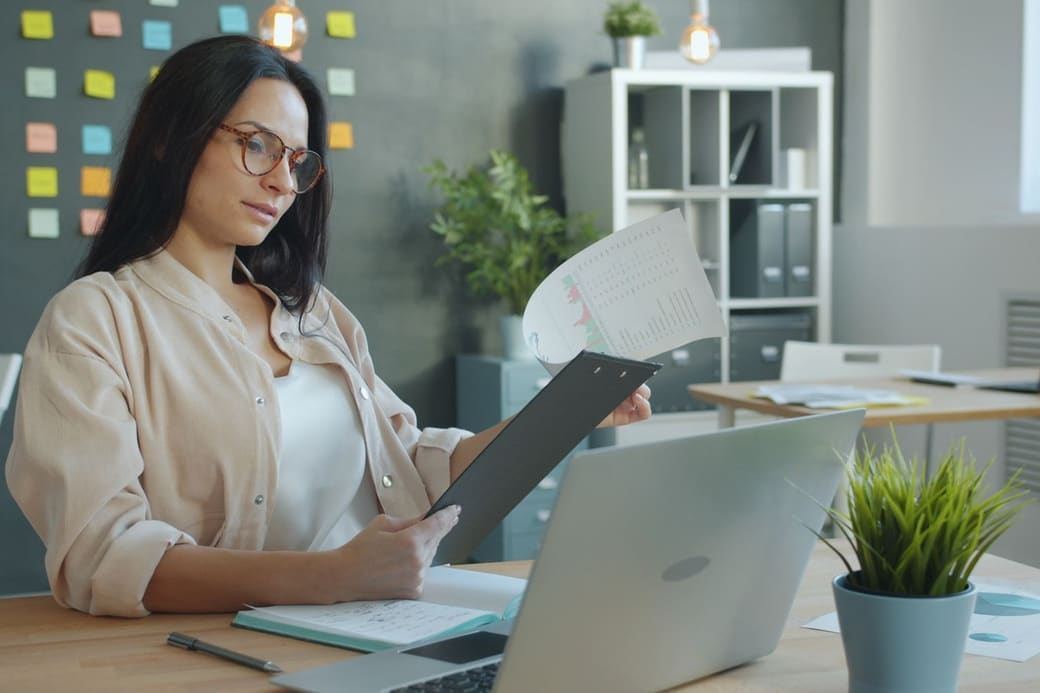 A woman at her office desk reading a document on a clipboard beside an open laptop