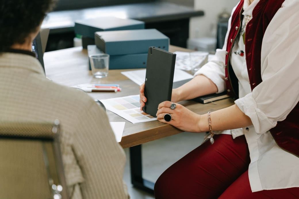 Two people across a meeting table covered with printed specifications and product samples