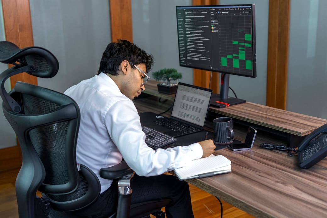 An analyst in a white shirt working at a desk with a laptop and a monitor showing a data dashboard
