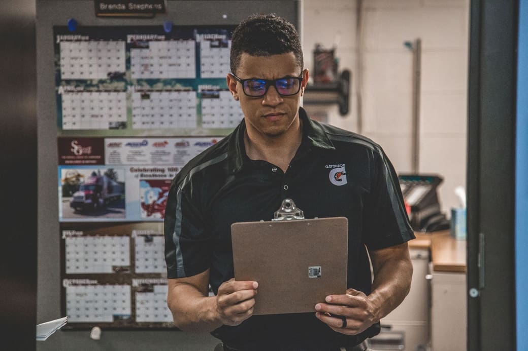 A warehouse supervisor in a company polo reading a clipboard in front of a wall of shift calendars