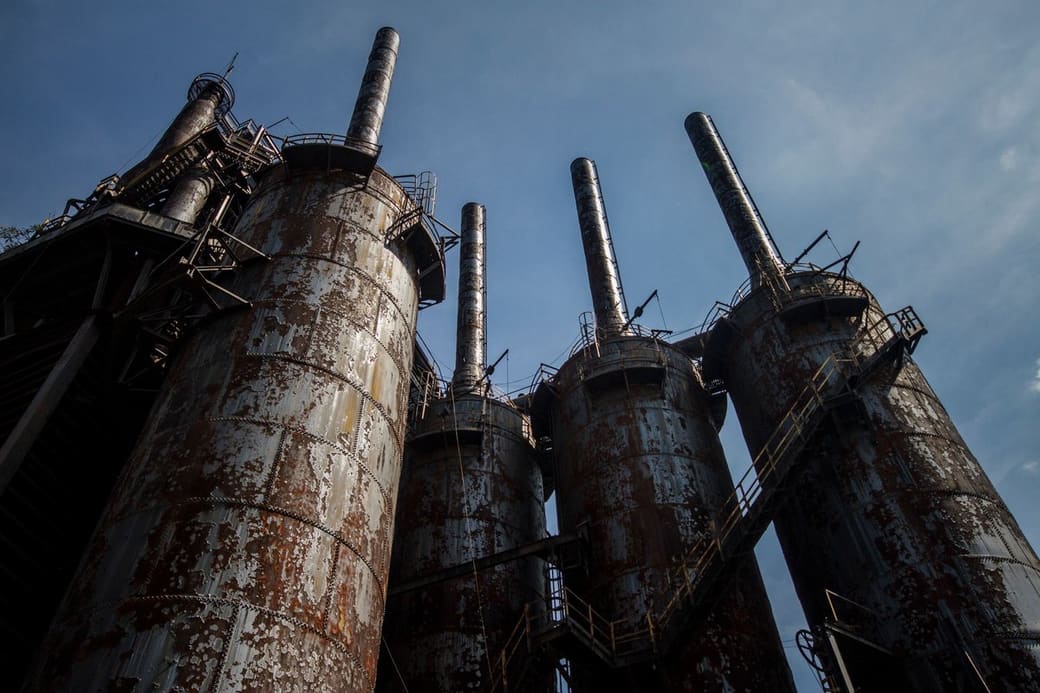 Looking up at a row of massive rusted steel blast furnaces and tall stacks from a former steel mill, with catwalks and stairways against a blue evening sky.