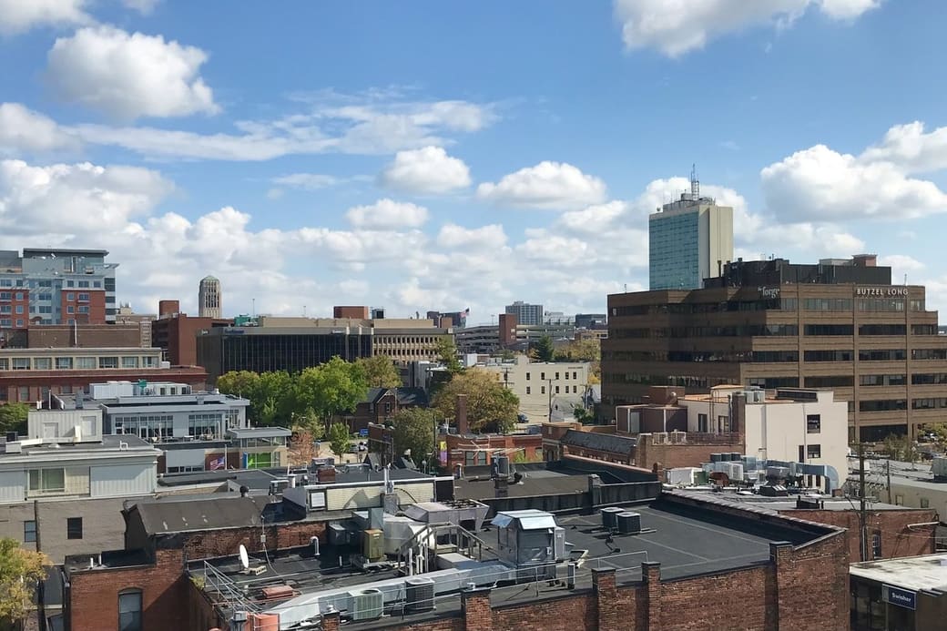 Rooftop view across a small college-town downtown of brick and mid-rise buildings, with one slender blue-glass tower rising above them under puffy white clouds.