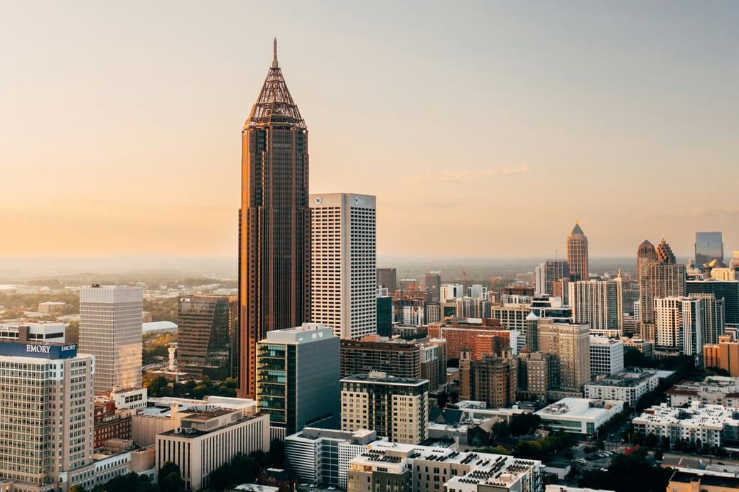 A dark tower with a latticed spire standing above a dense downtown at sunset, the city fading into haze behind