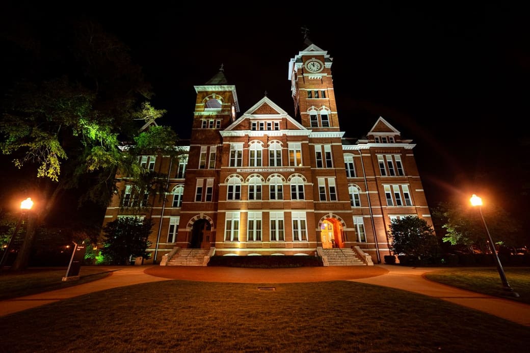 A red-brick Victorian hall with a white-trimmed clock tower, lit warmly at night, stands behind a curved lawn with glowing lamp posts and dark trees.