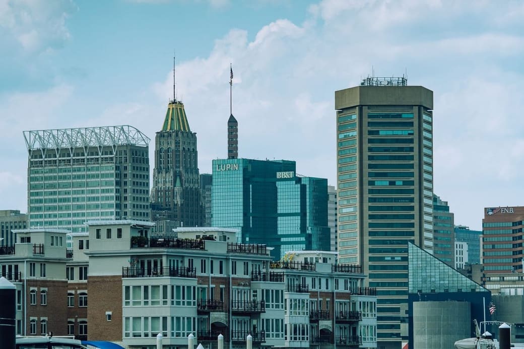 A daylight city skyline of glass and stone towers, one topped in gold, rising behind a row of waterfront apartment buildings with balconies
