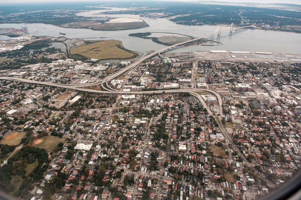 Aerial view over a low-rise coastal city of tree-lined streets, with elevated highway ramps curving toward a white cable-stayed bridge spanning a wide harbor and container port beyond.
