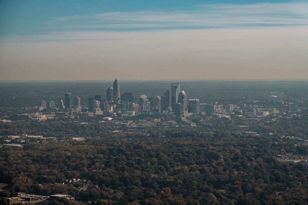 A cluster of downtown towers seen from far above across a wide canopy of trees, under a hazy sky