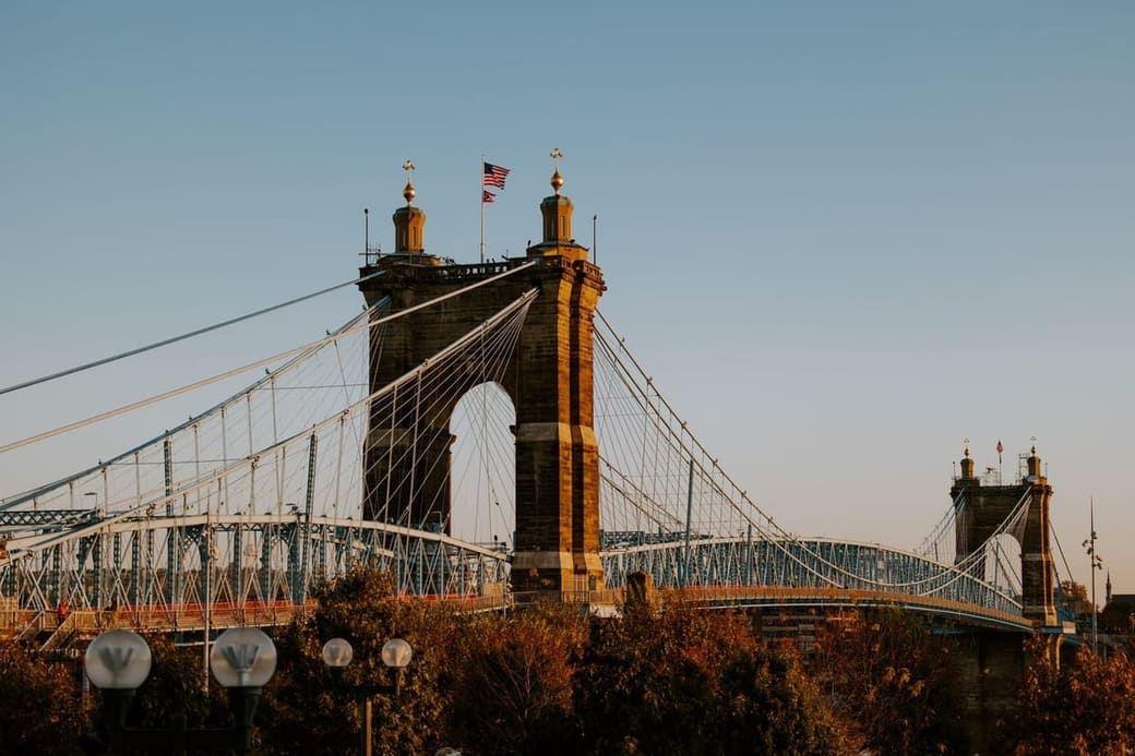 The stone towers of a suspension bridge lit low and gold, cables fanning down to a blue steel deck, with autumn trees along the bottom of the frame