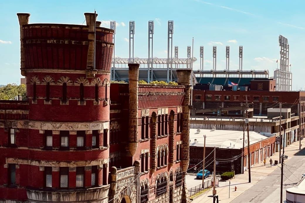 A red brick armoury with a rounded corner turret in the foreground and the floodlight masts of a baseball stadium rising behind it, seen from above a quiet street