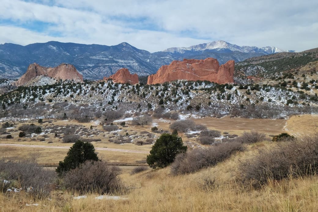 Jagged red sandstone rock formations rise from dry golden grassland dotted with juniper and light snow, with a snow-capped mountain peak and forested ridges behind.