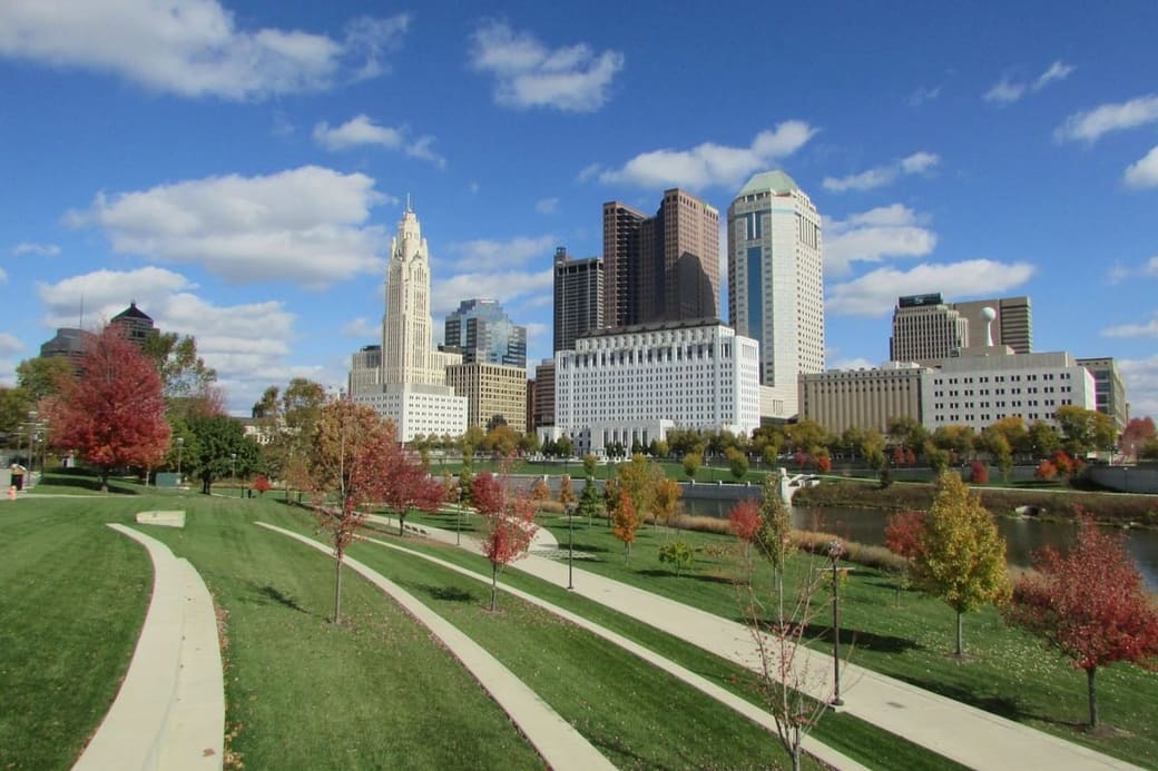 Parkland with curving paths and young trees in autumn color, a downtown skyline with a white spired tower rising behind it