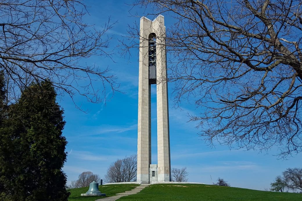A tall pale stone bell tower with an open arched top full of bells rises from a grassy hill, framed by bare winter branches against a bright blue sky.