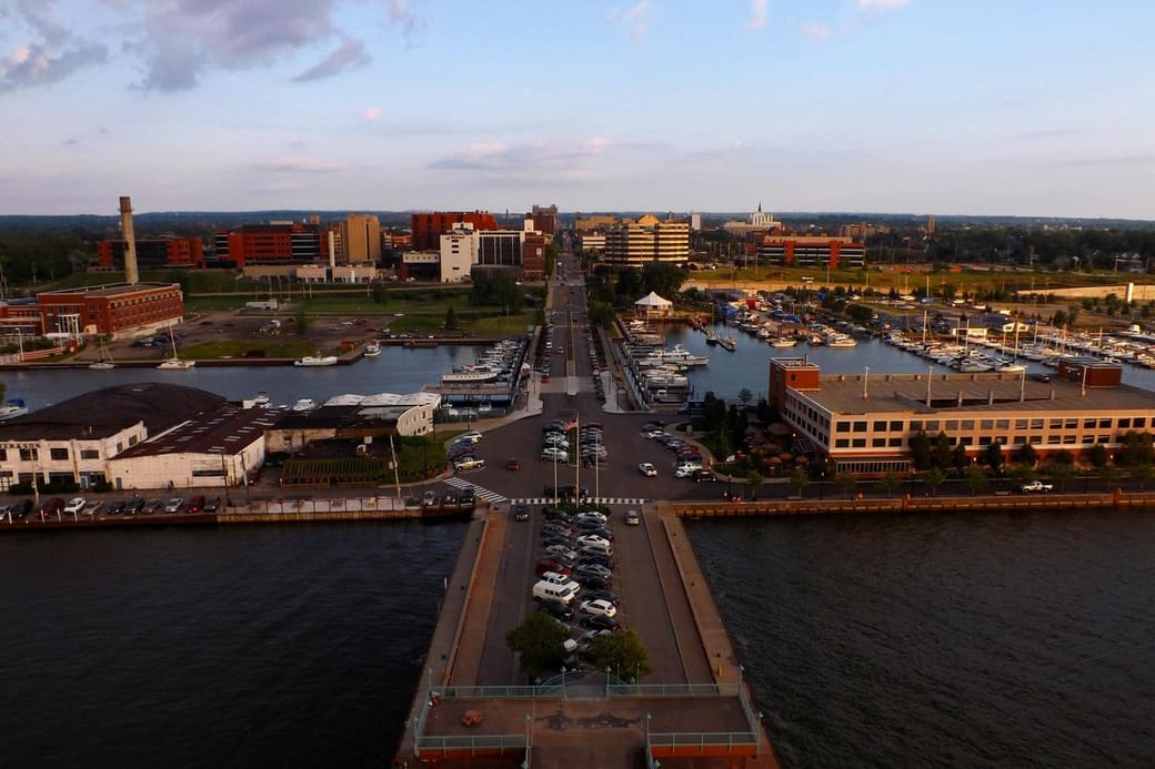 High view down a pier toward a lakeside downtown, with marinas full of boats on either side, a straight main street running inland and evening light on the buildings.