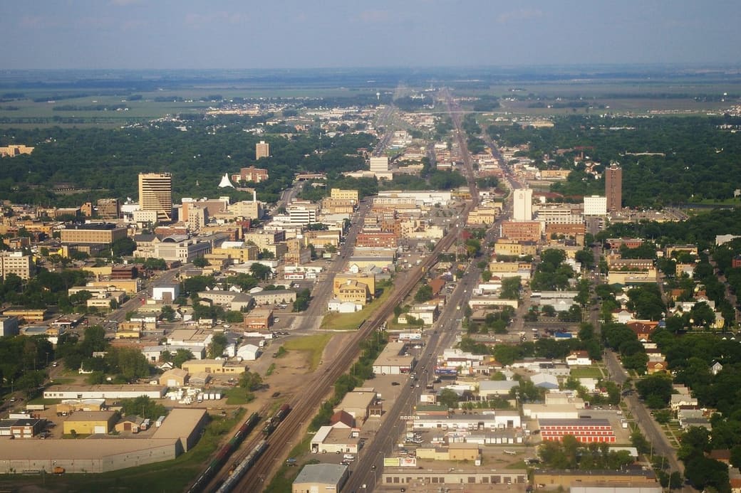 Aerial view of a small prairie city downtown, with a rail line and straight avenue running toward a flat green horizon past brick buildings and a few mid-rise towers.