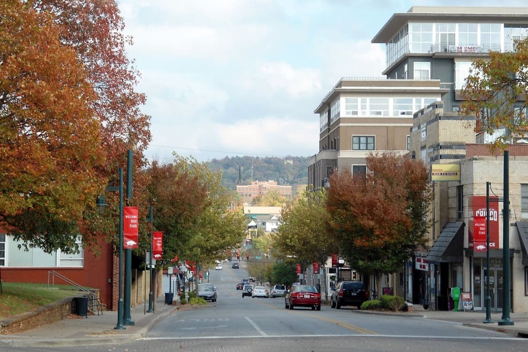 A downtown street in autumn slopes toward wooded hills, lined with orange-leaved trees, red street banners, shops and a modern mid-rise building on the right.