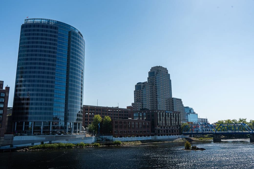 A curved glass tower and older brick and stone buildings along a wide river, a blue steel bridge at the right