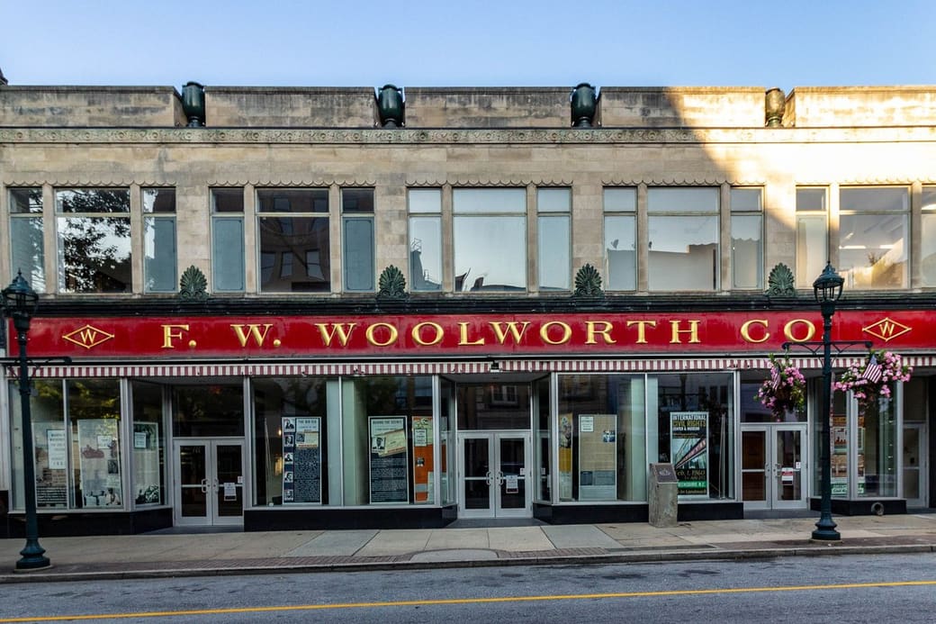 A two-story pale stone commercial building on a quiet downtown street, with a long red storefront sign in gold lettering, tall windows and black lampposts under a clear sky.