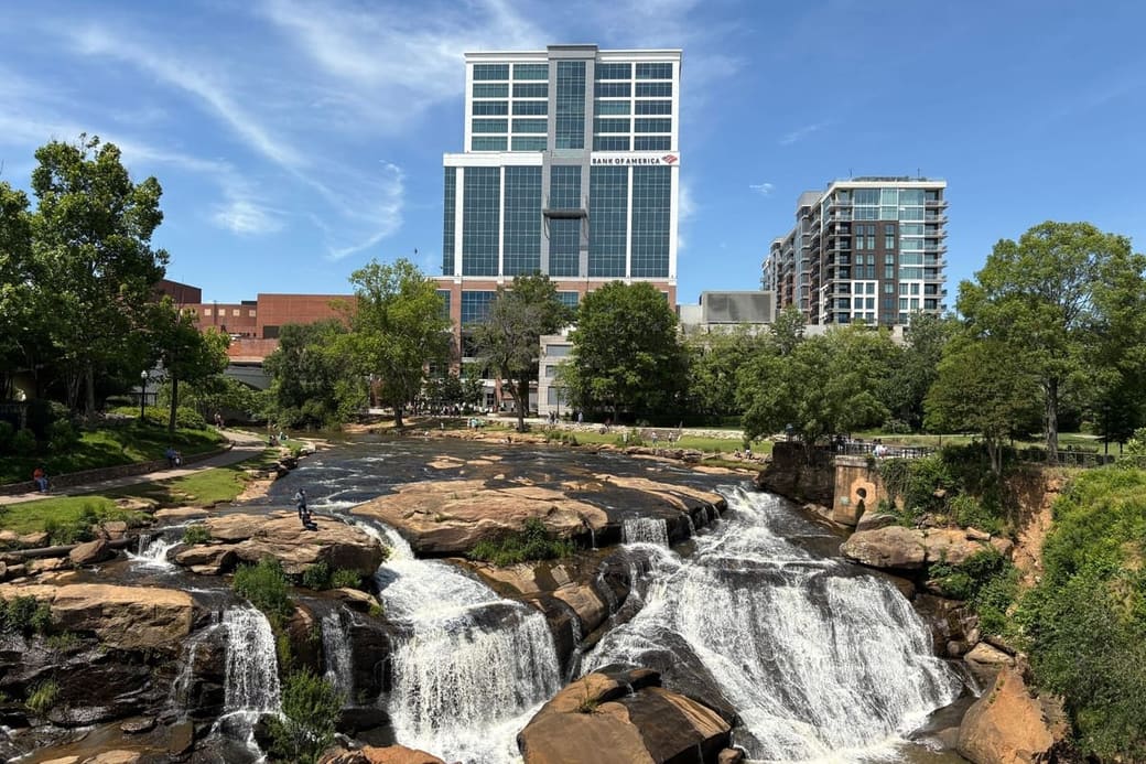 A wide rocky waterfall running through a park in daylight, with modern apartment blocks and trees along the bank above it
