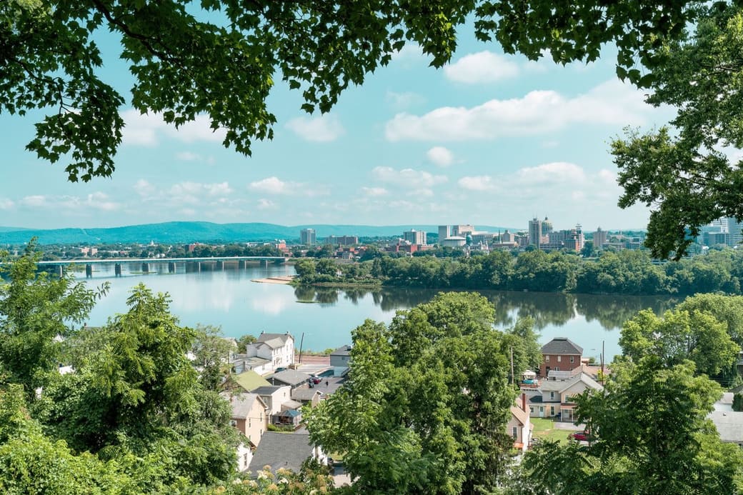 A small downtown skyline and long low bridge sit across a broad, calm river, seen from a leafy hillside above houses, with blue ridges on the horizon on a summer day.
