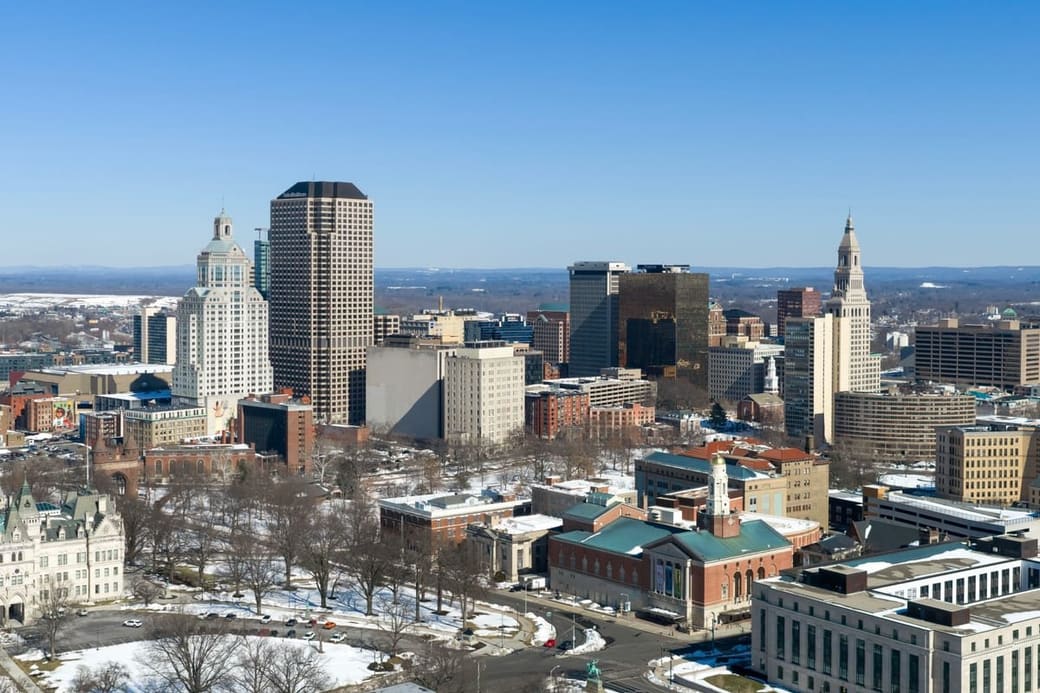 A winter cityscape seen from above, a gold-domed building among low roofs in the foreground and a cluster of dark office towers behind under a pale sky