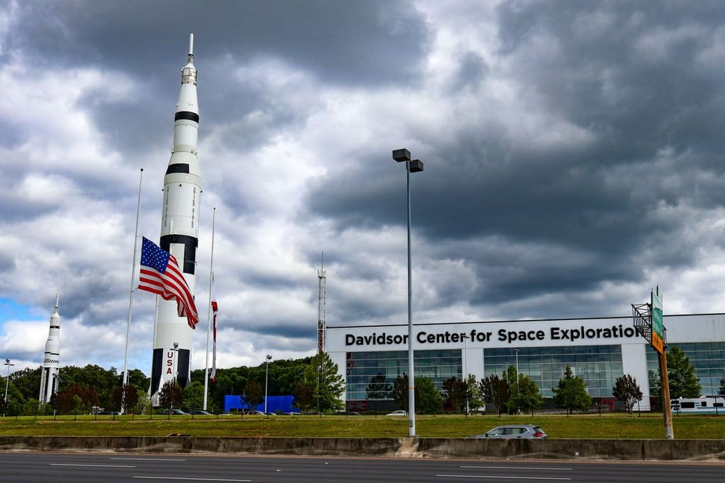 A towering white Saturn-style rocket and a smaller rocket stand beside a long glass exhibition hall and an American flag, seen across a highway under heavy gray clouds.