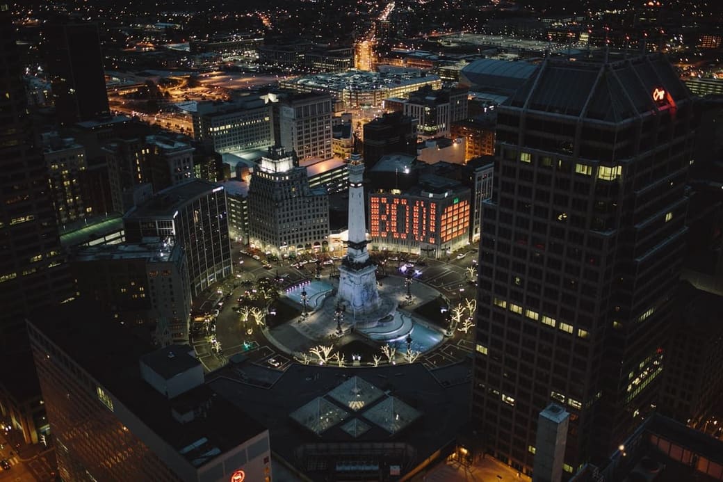 A night aerial of a circular plaza with a lit stone monument at its center, ringed by office towers and streets picked out in light
