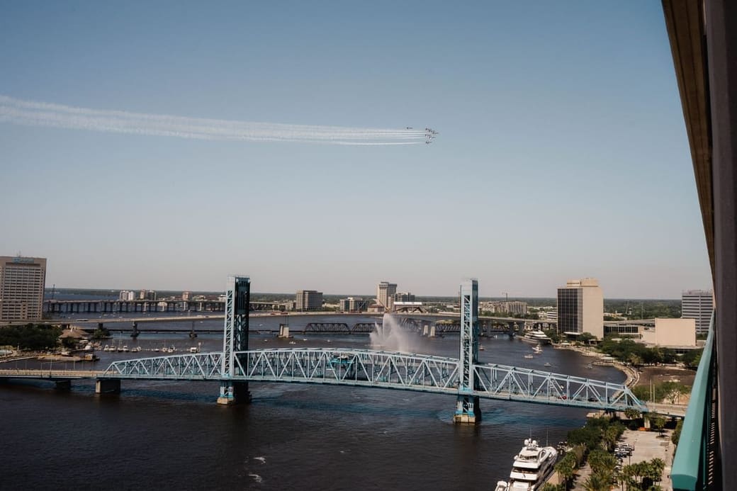 A blue steel lift bridge crossing a wide river under a clear sky, with a small cluster of downtown towers beyond it