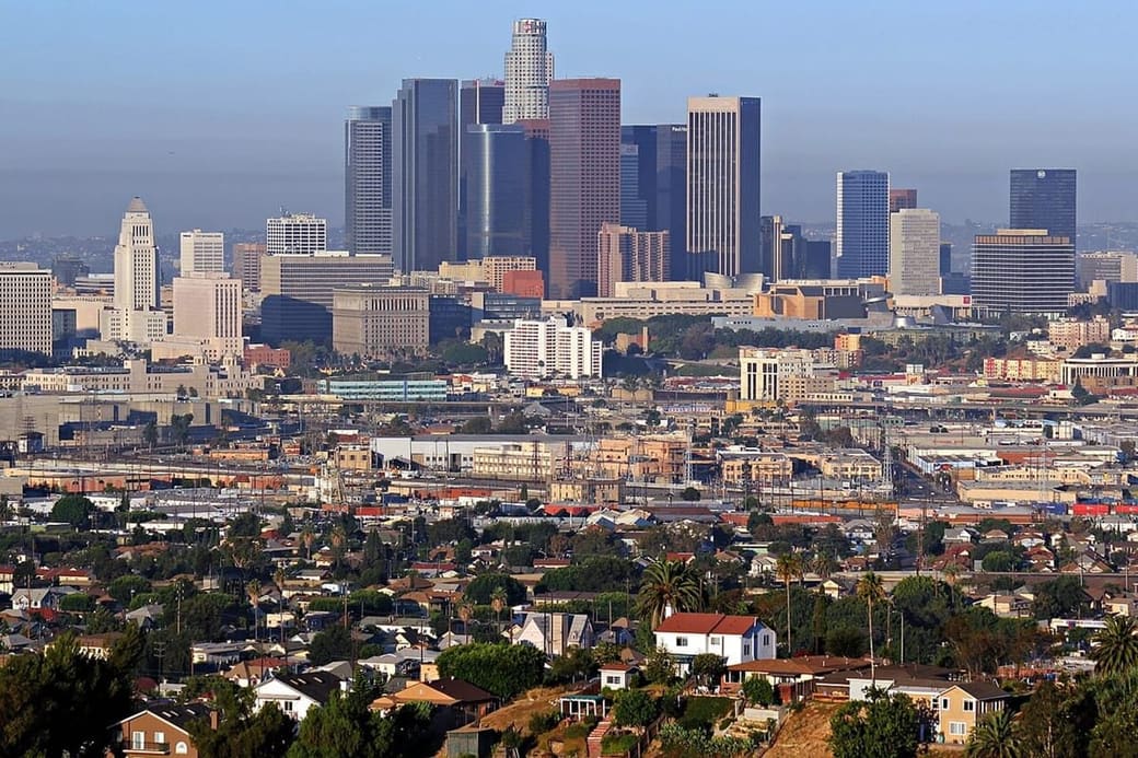 A downtown cluster of glass towers seen from a hillside, with low houses and palm trees filling the foreground