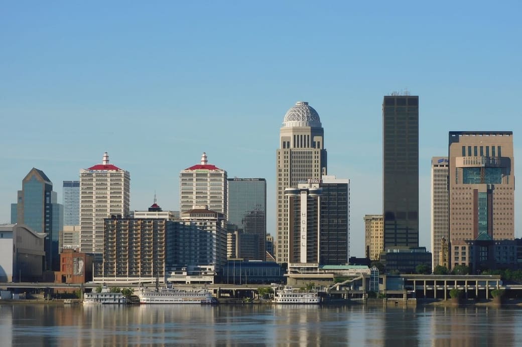 A downtown skyline seen across a broad river in daylight, two towers with red-lit crowns standing out among the others