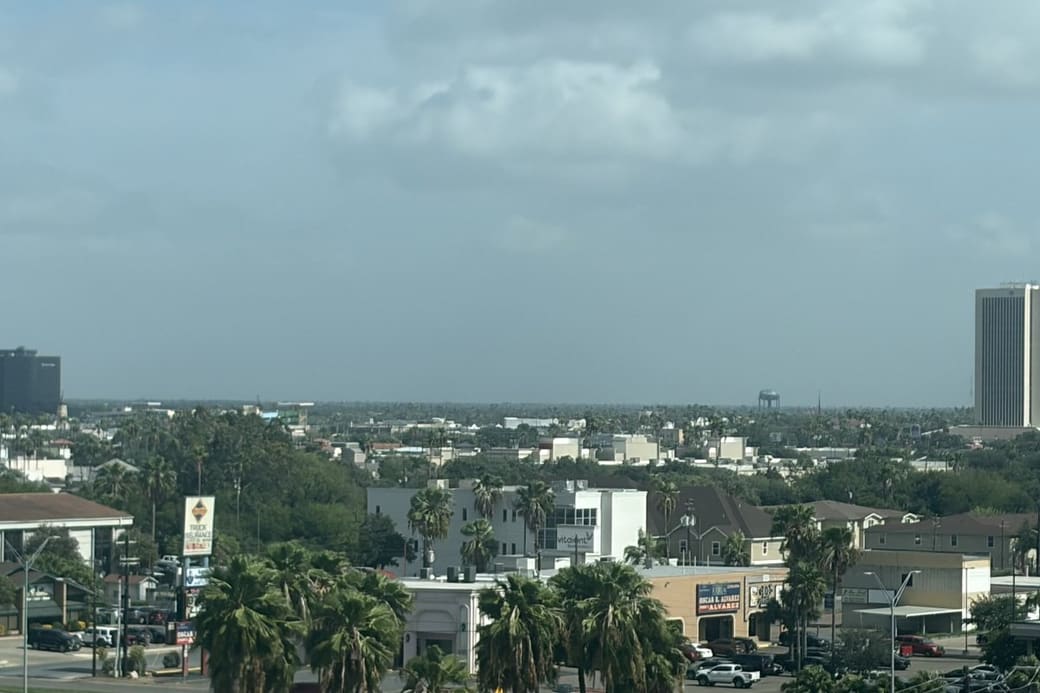 A flat, spread-out city of low buildings and palm trees under an overcast sky, with a single tall white office tower and a dark high-rise standing out on the horizon.
