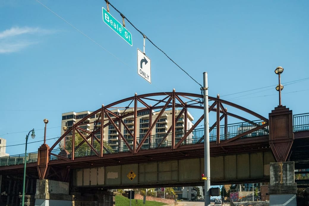 A steel arch bridge spanning a wide river under a clear blue sky, with a compact downtown of glass towers on the far bank