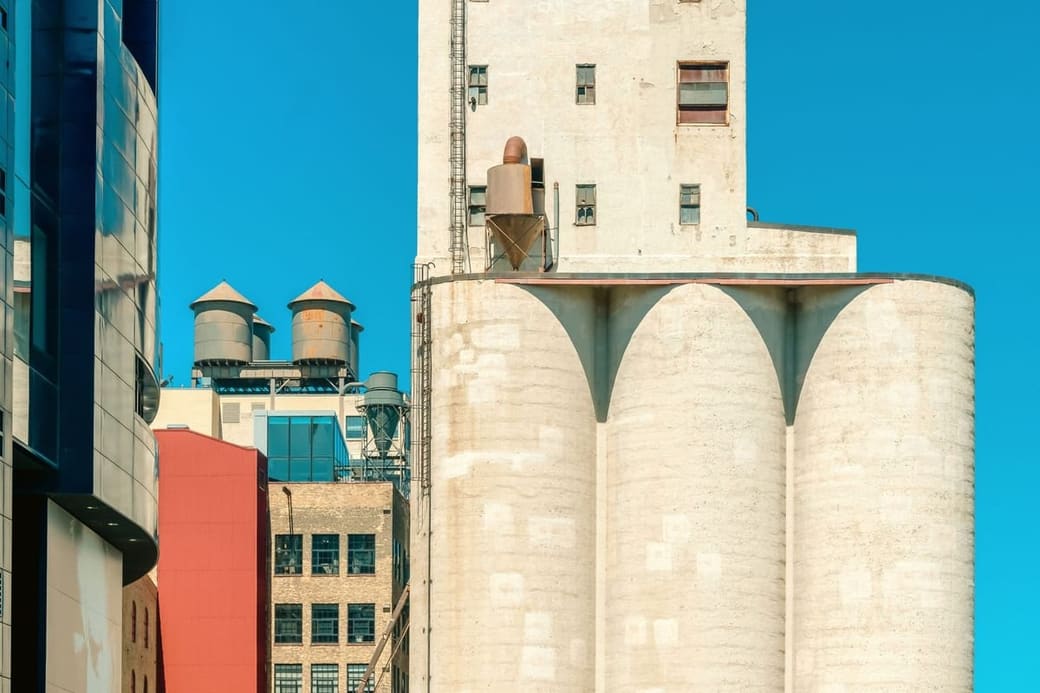 Concrete grain silos and a mill tower in pale stone beside a red brick warehouse and blue glass, under a hard blue sky