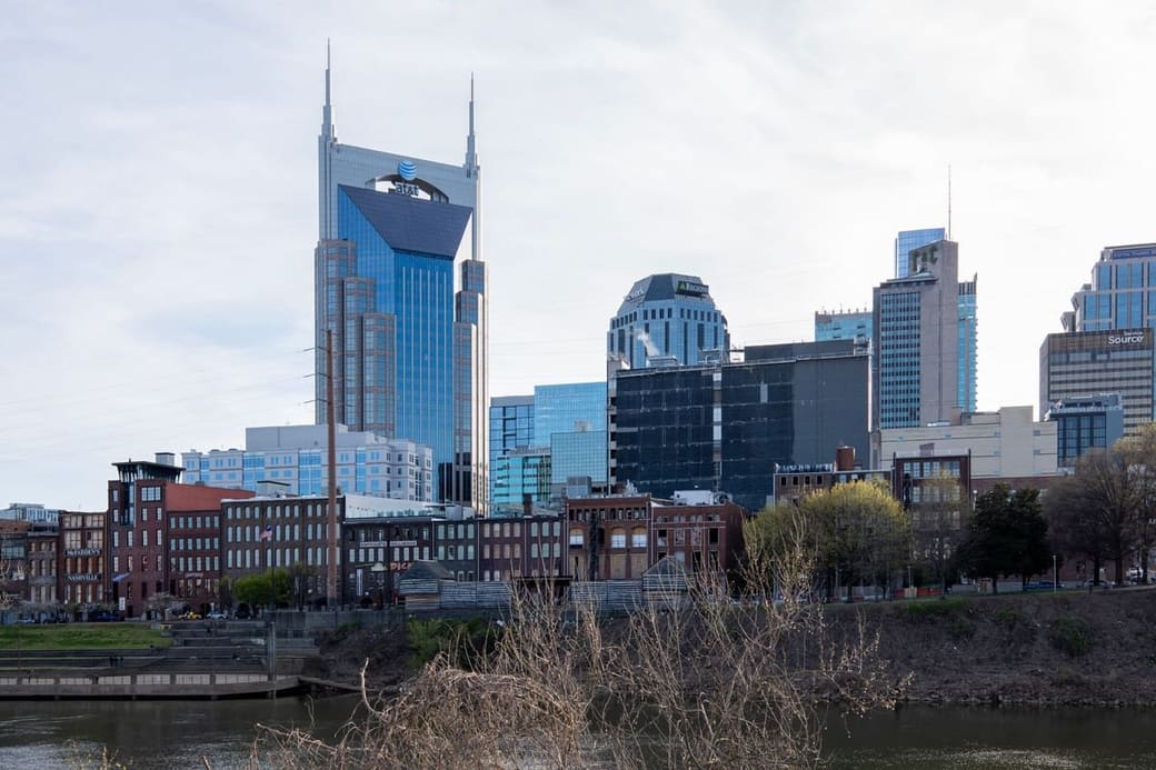 A city skyline across a river in flat daylight, a tall tower with two thin spires standing above the other buildings, low brick warehouses along the near bank