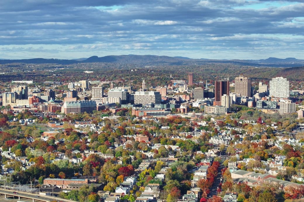 Elevated view over autumn-colored neighborhoods of wooden houses toward a compact cluster of downtown towers, with low wooded ridges and a cloud-streaked sky beyond.