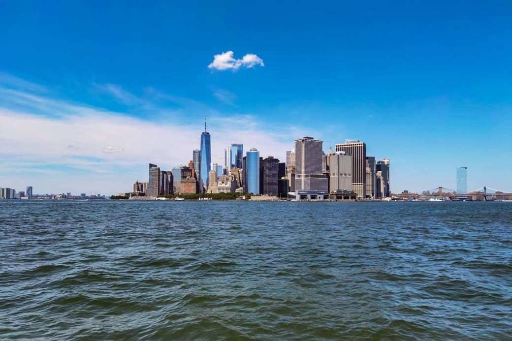 A dense cluster of towers on the tip of an island seen across open water under a deep blue sky, a suspension bridge at the right edge