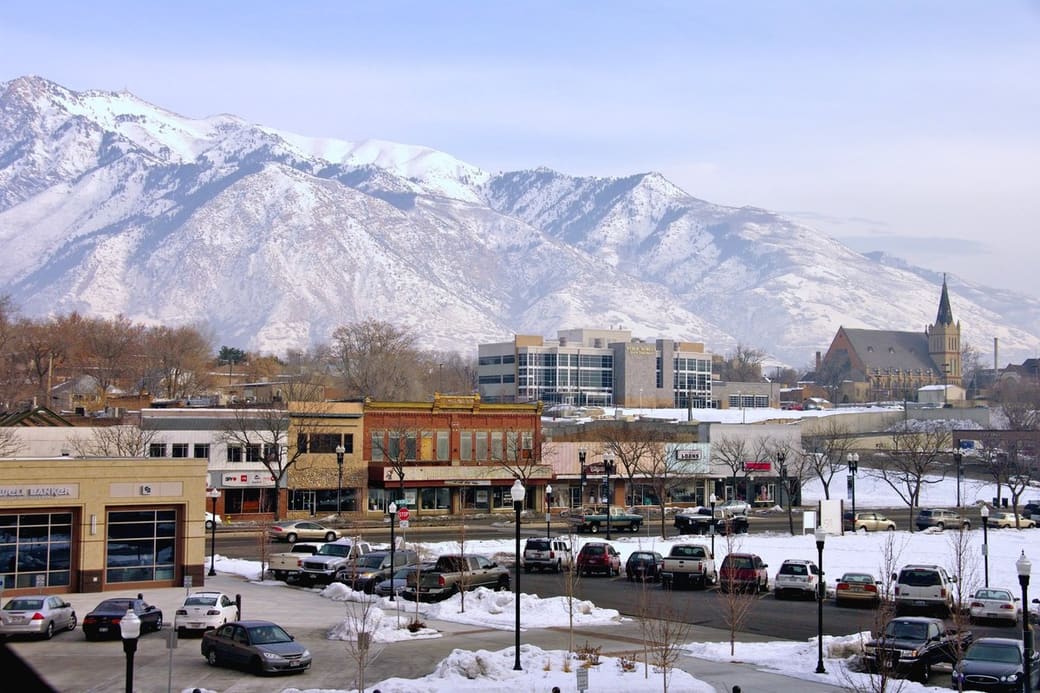 A small downtown of brick storefronts, a church spire and parked cars in winter, backed by a steep wall of snow-covered mountains under a pale blue sky.