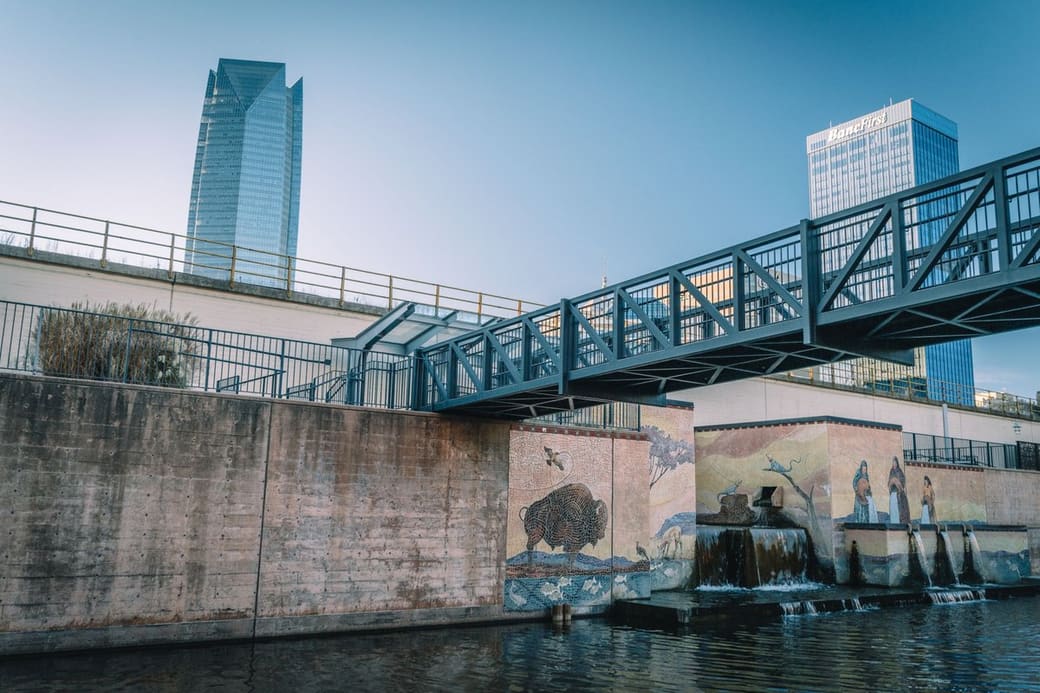 A canal lined with murals of bison and birds, a steel footbridge crossing overhead and two glass towers rising behind