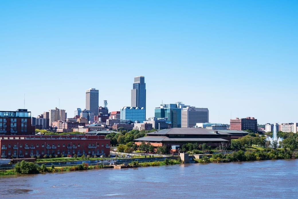 A downtown skyline across a wide brown river under a clear sky, with a red brick warehouse row and a fountain along the near bank