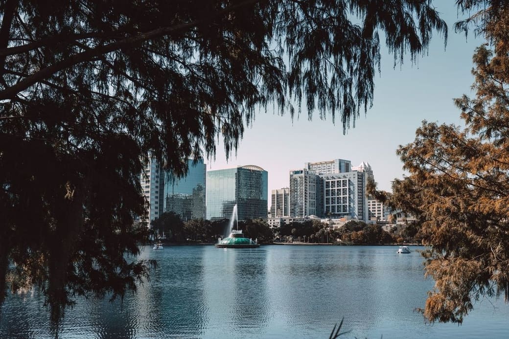 A lake with a fountain and a row of glass towers on the far shore, framed by dark overhanging branches