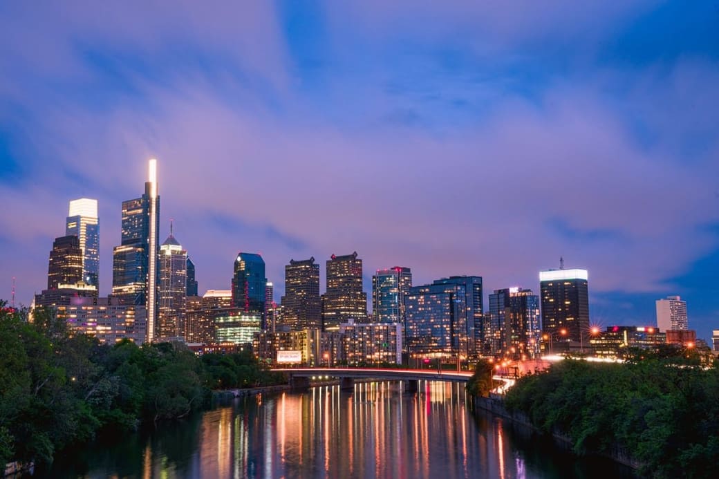 A city skyline lit at dusk under a violet sky, reflected in the river below with a bridge crossing in front of it