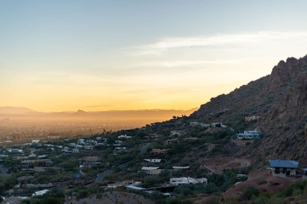 A hillside of low houses and desert scrub at sunset, a rocky ridge rising on the right and a hazy valley reaching to distant mountains