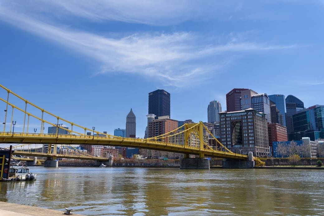 A yellow steel bridge crossing a wide river toward a compact downtown of glass and brick towers under a clear sky