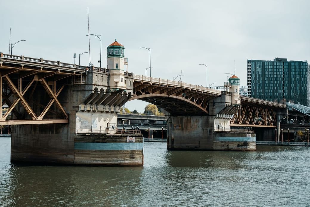 A dark steel truss bridge crossing a river in flat daylight, with low buildings along the far bank