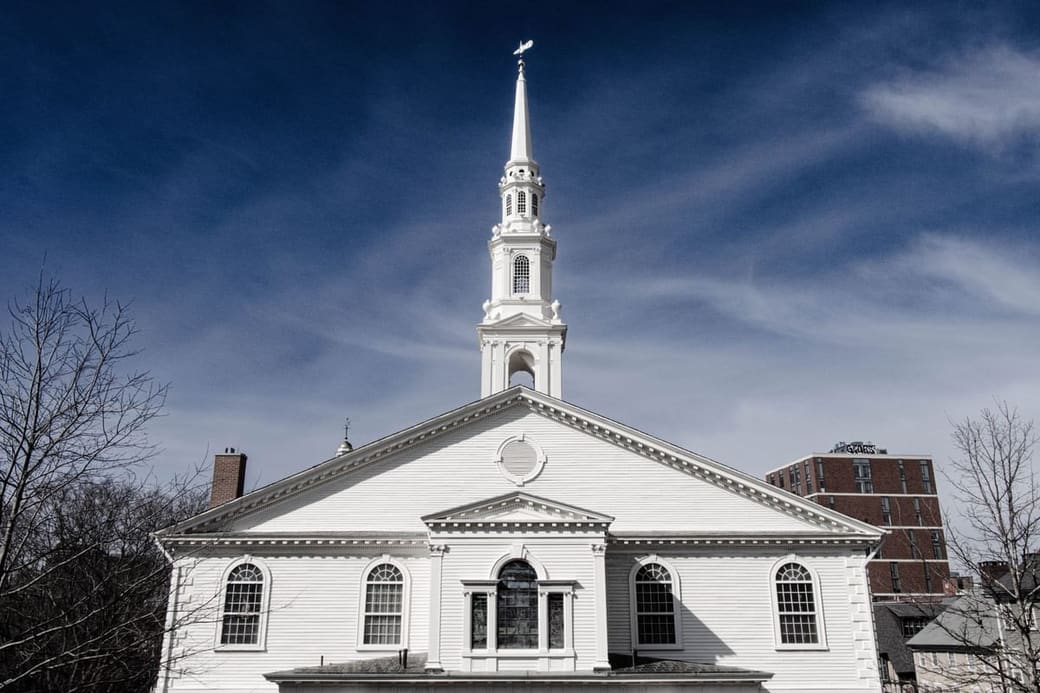 A white clapboard meeting house with a tall tiered steeple against a deep blue sky, bare winter branches at the edges