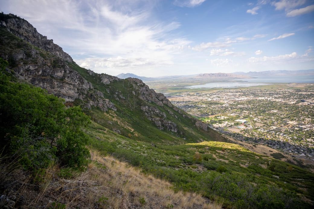 A steep green hillside falling away to a wide valley of towns and a lake, with a mountain range on the horizon
