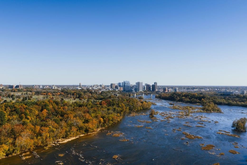 A wide rocky river through autumn woodland, a small downtown skyline on the horizon under a clear blue sky