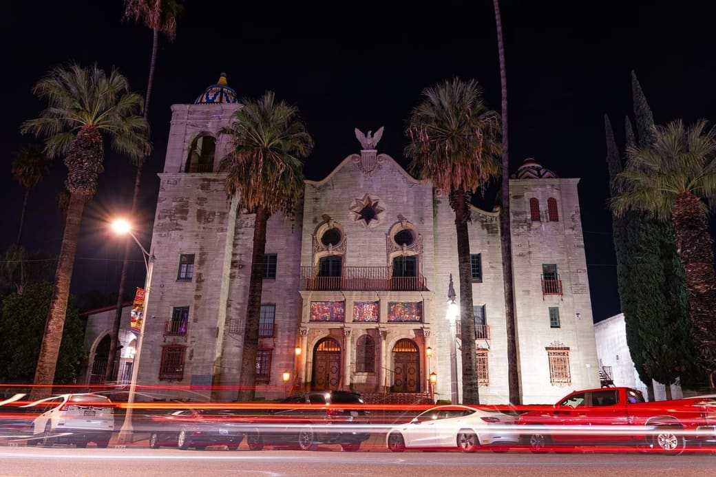 A floodlit stone mission-style building with arches and palm trees at night, light trails from passing cars along the street in front