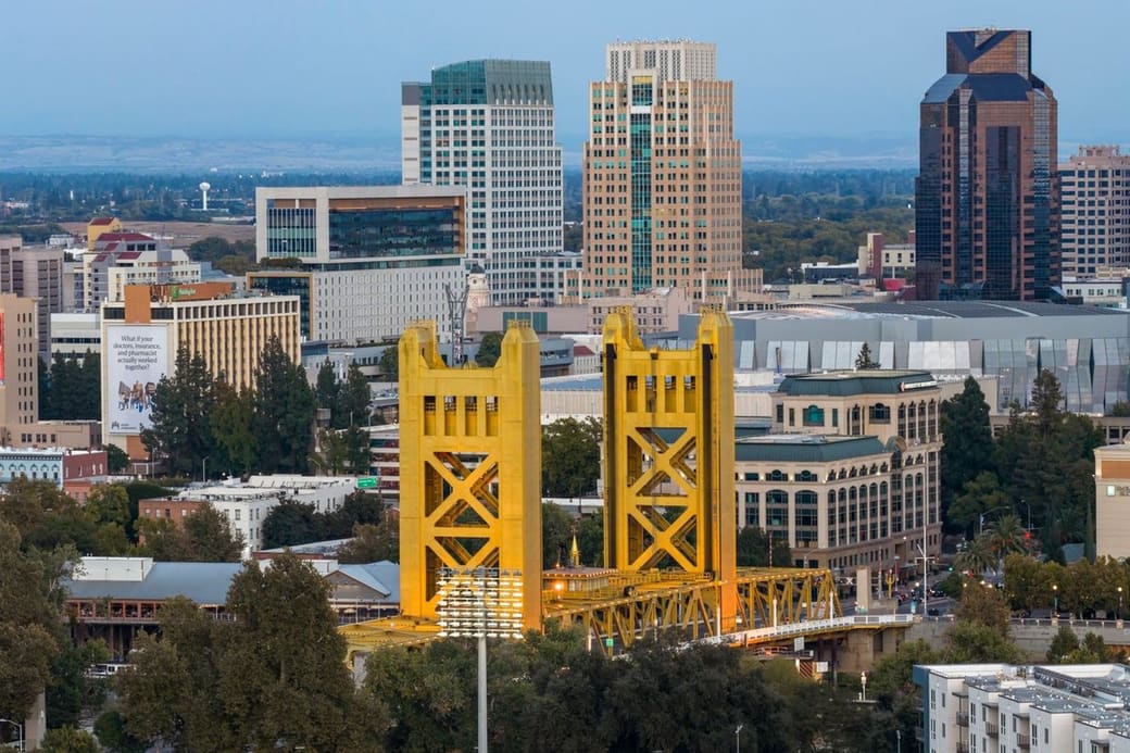 A gold lift bridge with twin towers in front of a compact downtown of office towers, trees along the riverbank